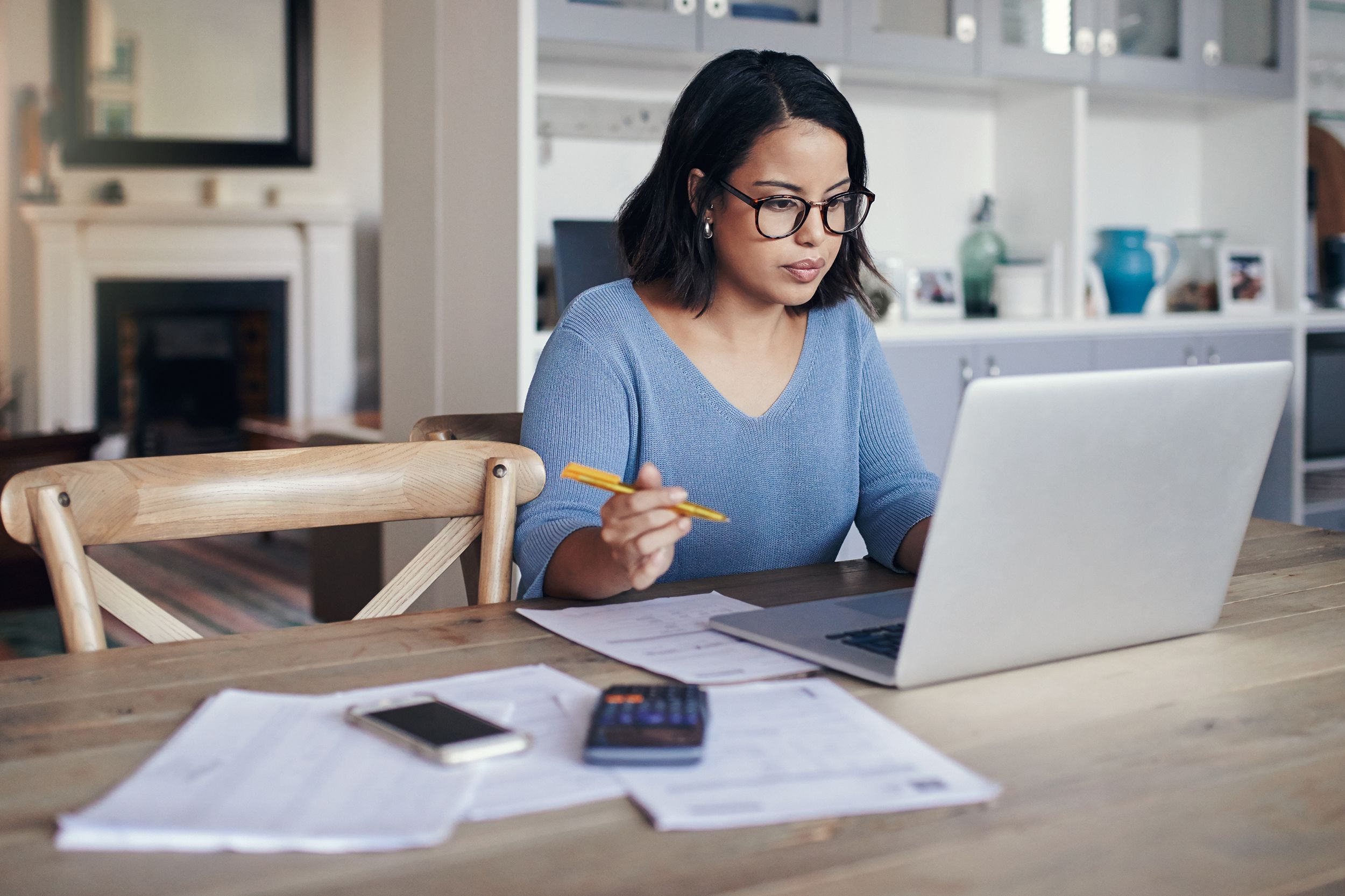 woman at home looking at laptop with papers and calculator on desk