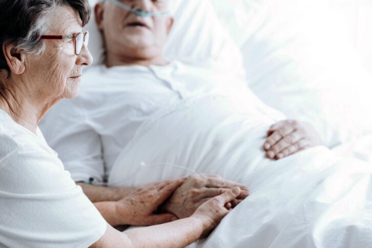 elderly woman holding husband's hand in hospital bed