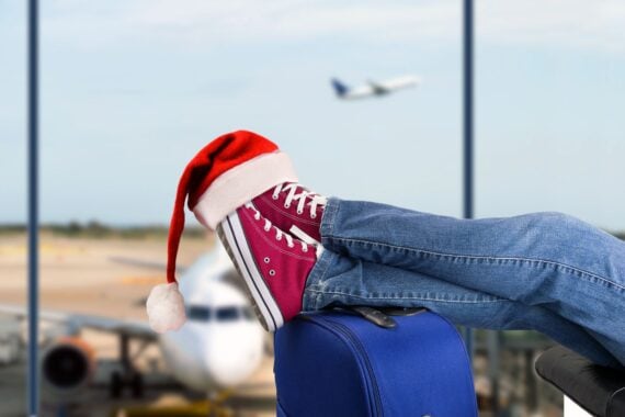close-up of feet at airport gate sitting on suitcase with santa hat hanging from one shoe, with airplane in the background
