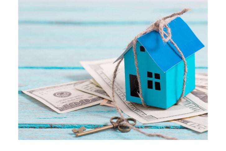 Paper house with key wrapped around it with money on a blue wooden background