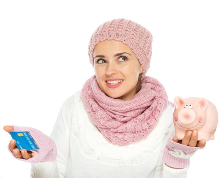 confused woman in knit winter clothing holding credit card and piggy bank
