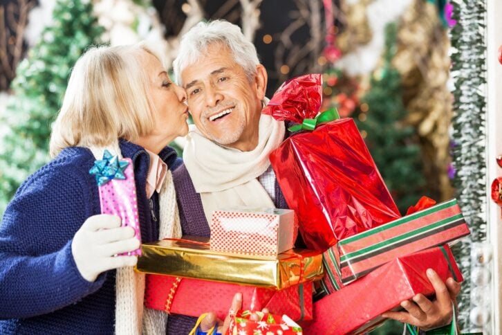 senior woman kissing man holding stacked Christmas presents at store
