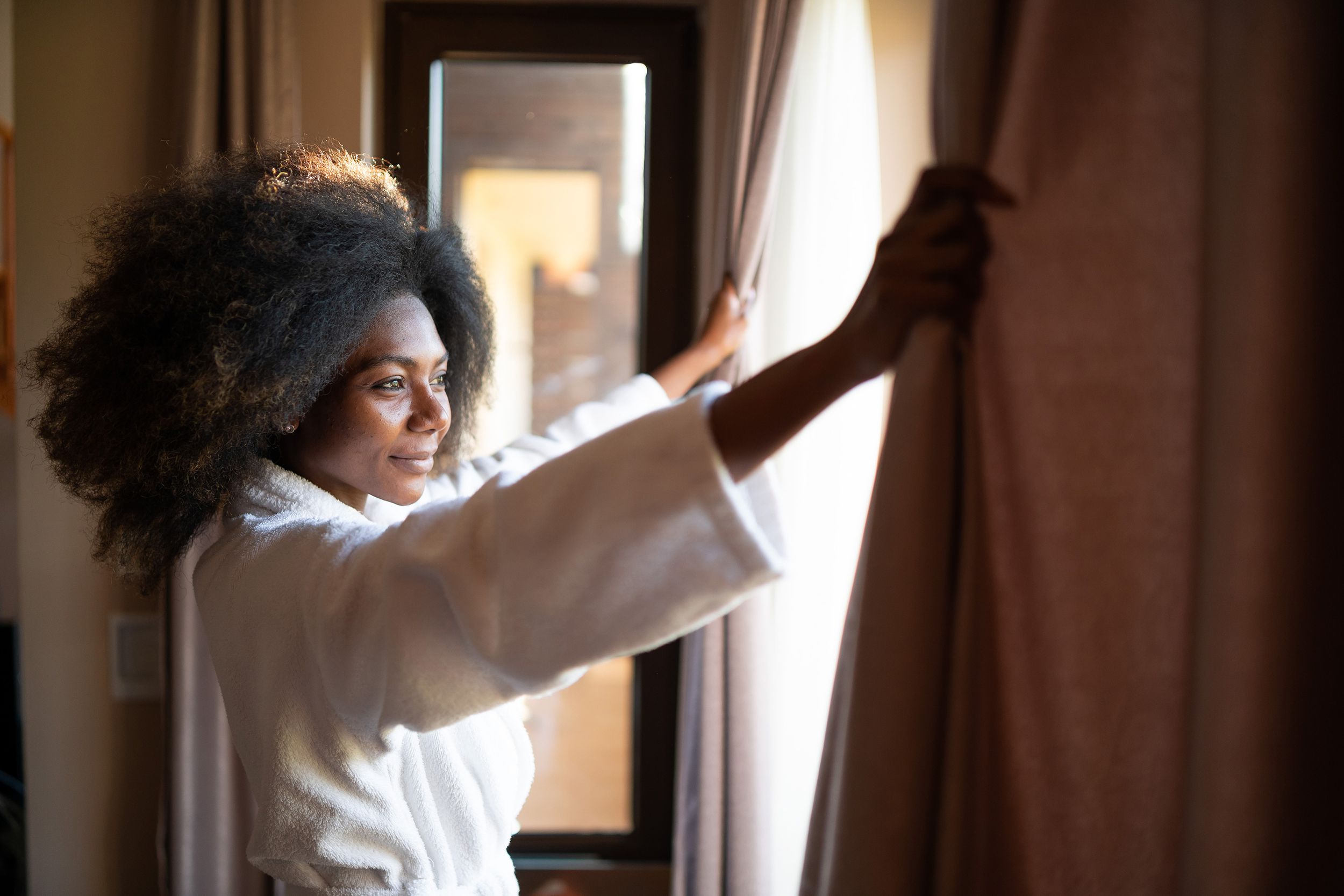 African woman pulling the curtains in hotel room, in the morning