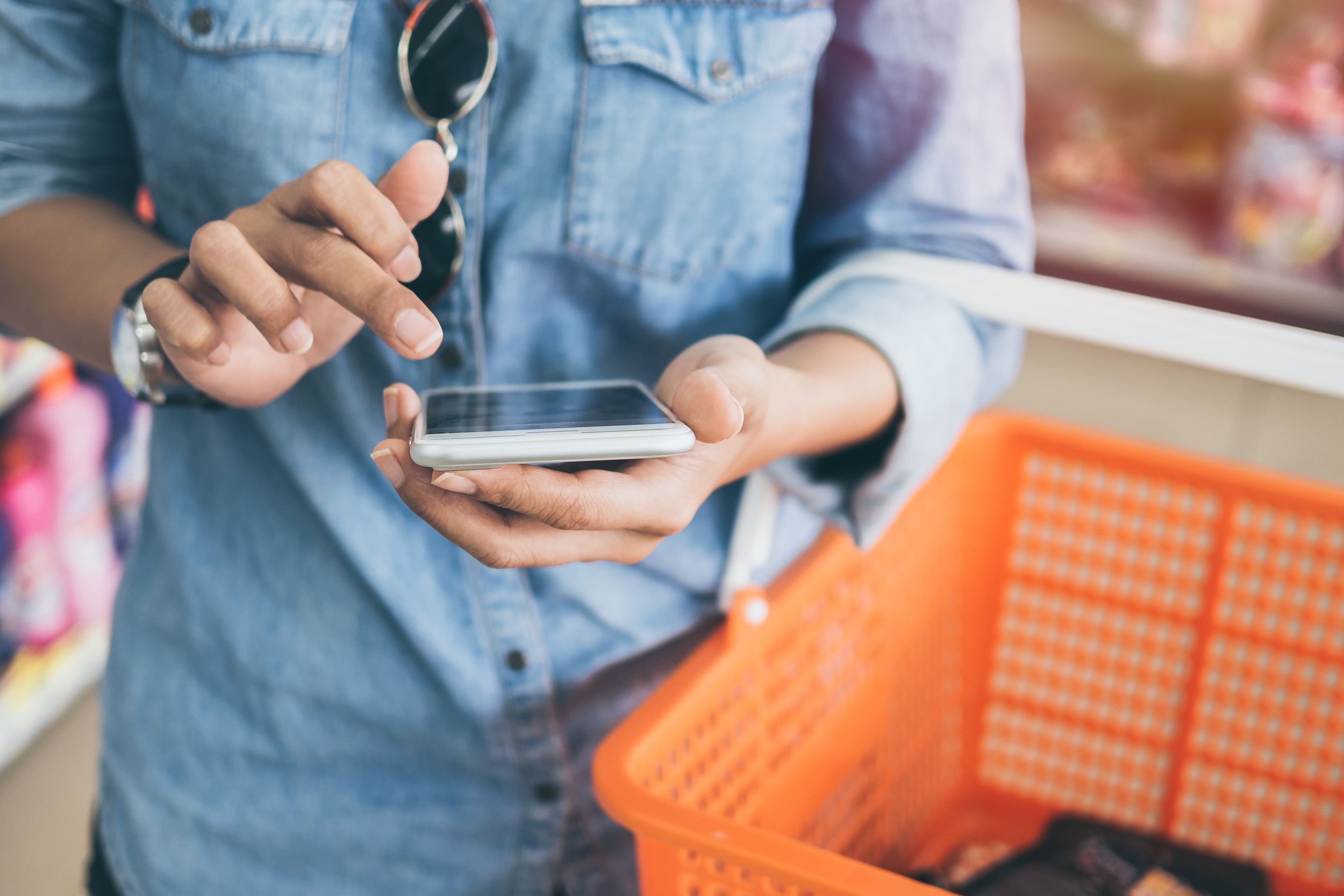 woman wearing blue jeans shirt and sunglasses using mobile phone to compare price and holding orange shopping basket in mini mart background