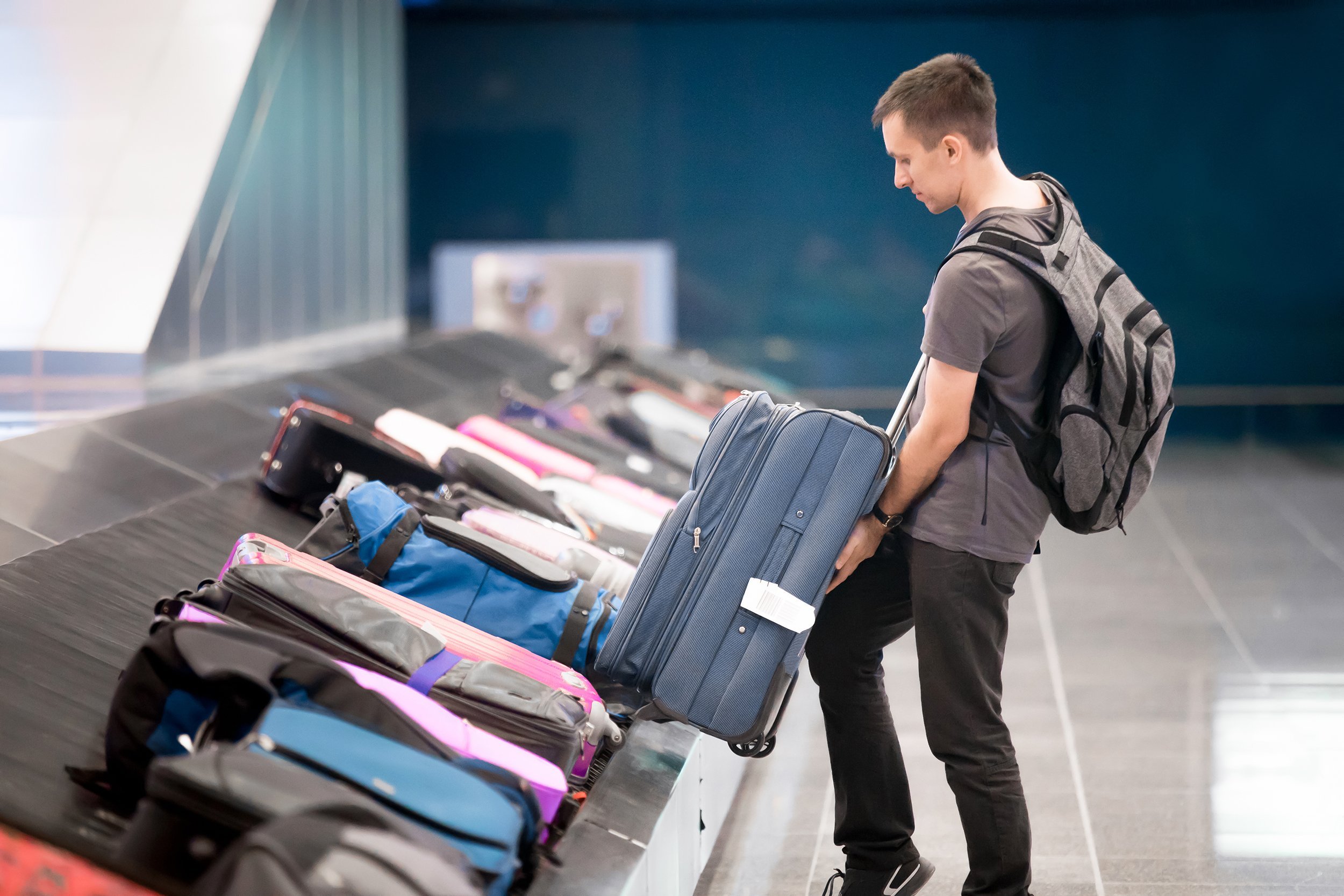young man passenger with carry-on backpack collecting his luggage at conveyor belt in arrivals lounge of airport terminal building
