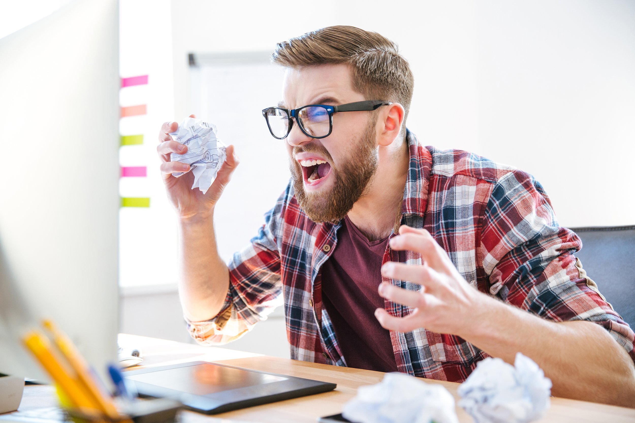 angry crazy modern designer in glasses with beard yelling and crumpling paper on his workplace