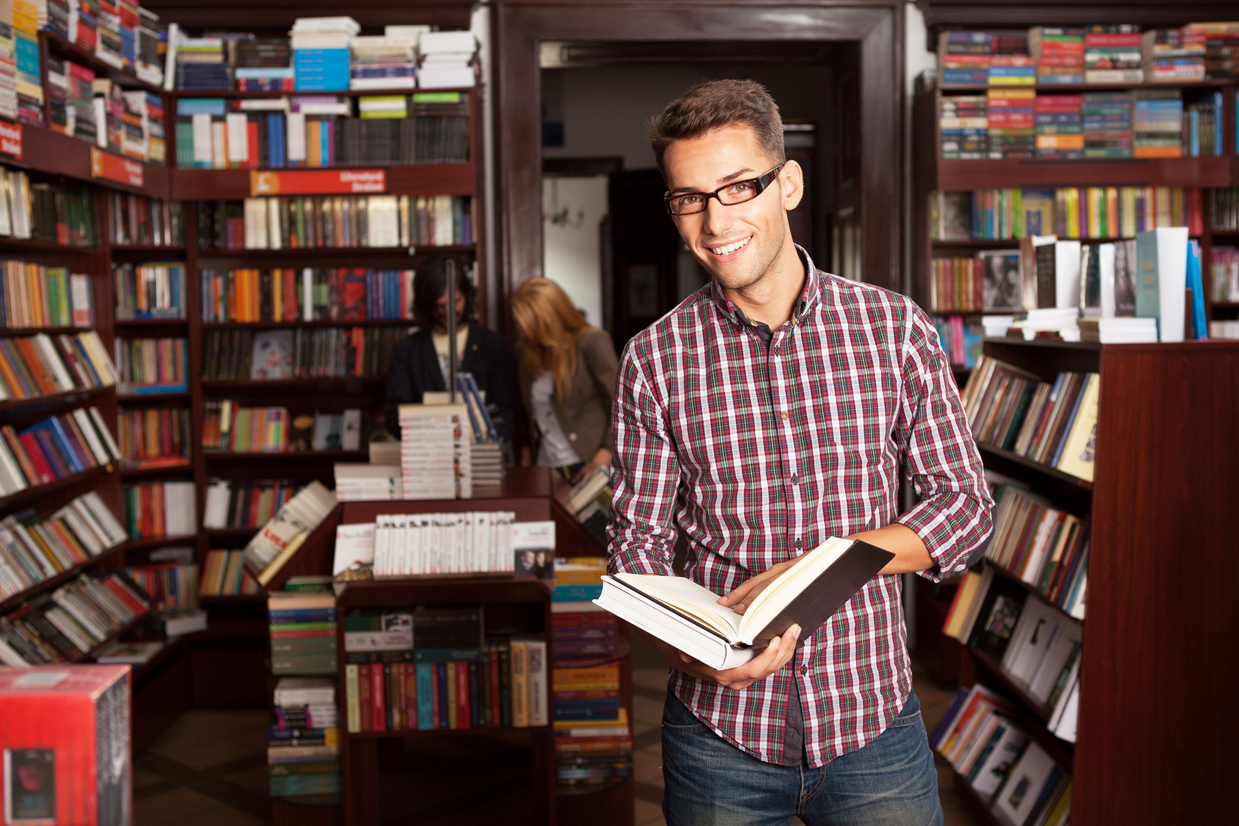 man with eyeglasses in a bookstore with an opened book in his hands smiling