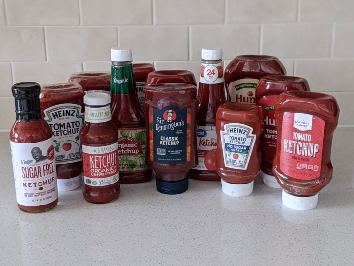 13 bottles of different ketchups all together on a speckled ivory kitchen countertop, light beige tile wall in the background