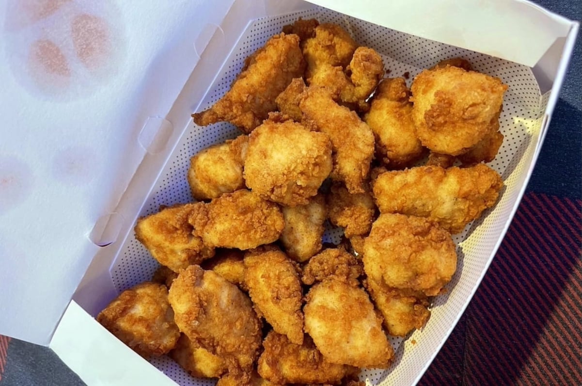 A white box filled with golden brown, crispy fried chicken nuggets—like those from fast-food chains with real chicken—sits open on a plaid surface.