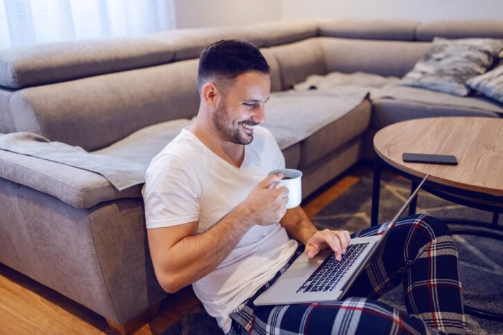 unshaven smiling man sitting on floor in living room, surfing on internet and drinking coffee in morning