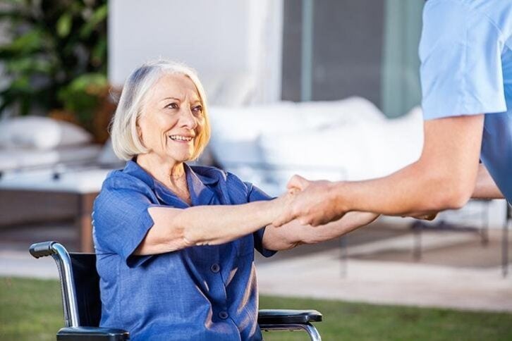 male nurse helping senior woman to get up from wheelchair at nursing home