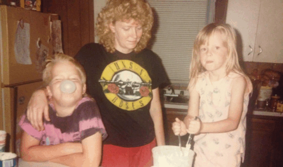 A woman stands in a kitchen with two children; one blows a bubble with gum while the other stirs something in a bowl. The woman wears a Guns N' Roses shirt and smiles at the children.