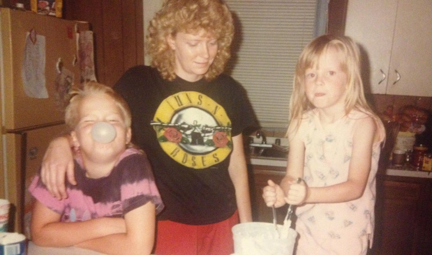 A woman stands in a kitchen with two children; one blows a bubble with gum while the other stirs something in a bowl. The woman wears a Guns N' Roses shirt and smiles at the children.