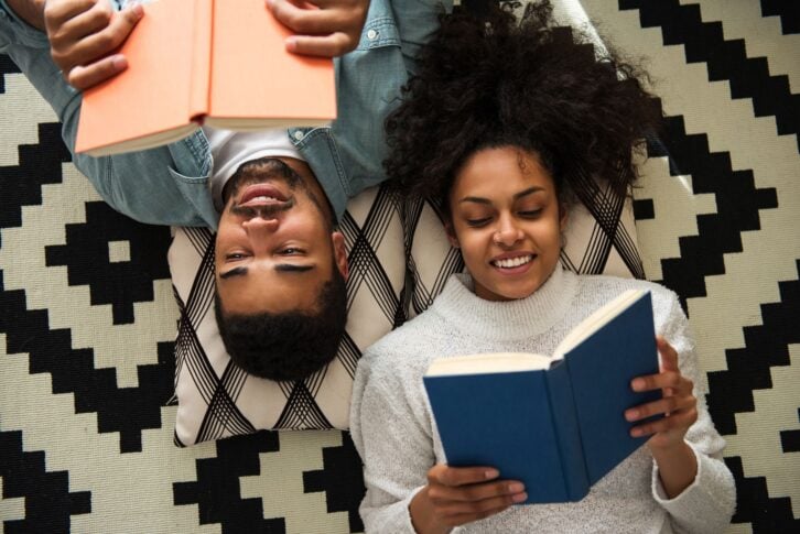 A male and a female laying on the floor, facing up, each holding and reading a book, male is upside down in image, on a geometric white and black carpet