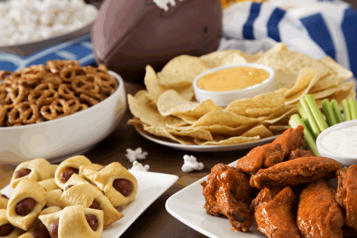 A selection of tailgating snacks including hot wings, pigs in a blanket, queso, and chips with a football in the background