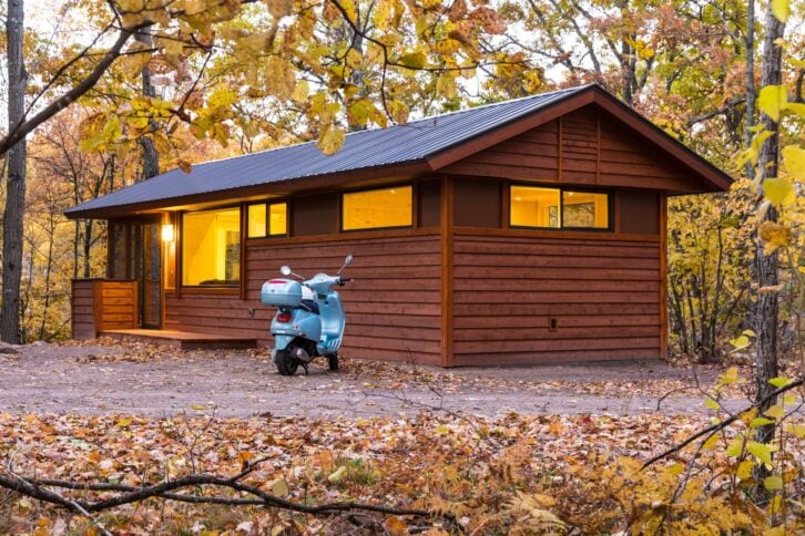 A tiny home in Canoe Bay Escape Village, Rice Lake, Wisconsin, leaves on yard and framed by a tree in the foreground during autumn, a light blue moped parked in front of it
