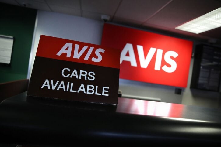 Avis 'Cars Available' sign on desk in foreground, red Avis sign slightly blurred in the background, slight diagonal