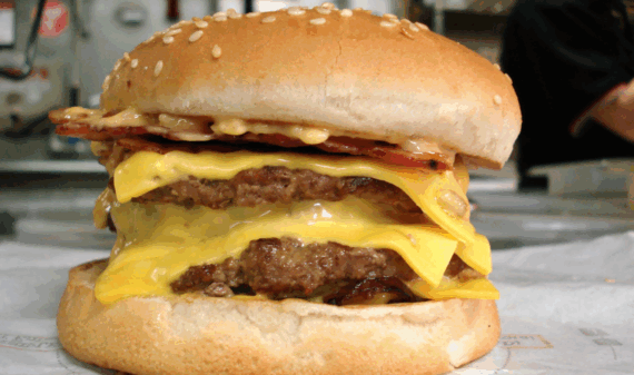 A close-up of a double cheeseburger with two beef patties, melted cheese, crispy bacon, and a sesame seed bun. The burger is on a sheet of paper, with a blurred kitchen background.