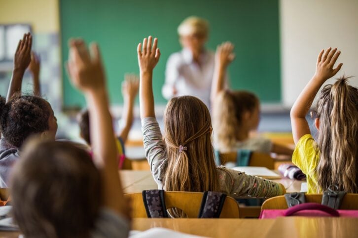 Back view of elementary students raising their arms while in class, four rows of student sitting at their desks, blurred teacher at the front of the classroom