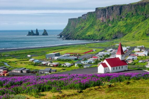 Basalt stacks and black sand beach of Reynisdrangar, South Iceland, vibrant green during summer, a small town and church