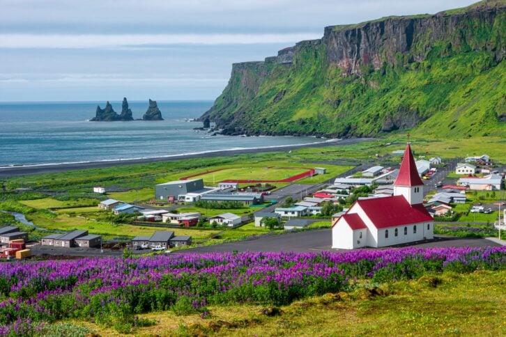 Basalt stacks and black sand beach of Reynisdrangar, South Iceland, vibrant green during summer, a small town and church