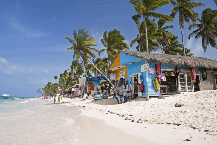Bavaro Beach, Punta Cana, Dominican Republic, tourists walking on the beach along colorful bungalows with souvenirs, palm trees and a bright blue sky in the background