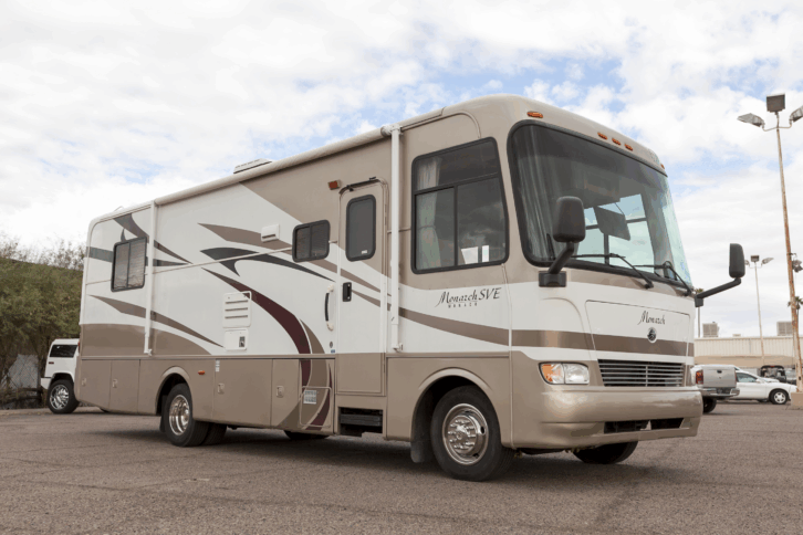 Side-view and slight front of a beige and white Monaco RV in Scottsdale, Arizona, on a parking lot, trees and building on the background left, cars in the parking lot, and white clouds in a blue sky
