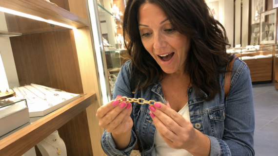A woman with dark hair and a denim jacket looks excitedly at a gold bracelet in a jewelry store, with display cases of jewelry in the background.