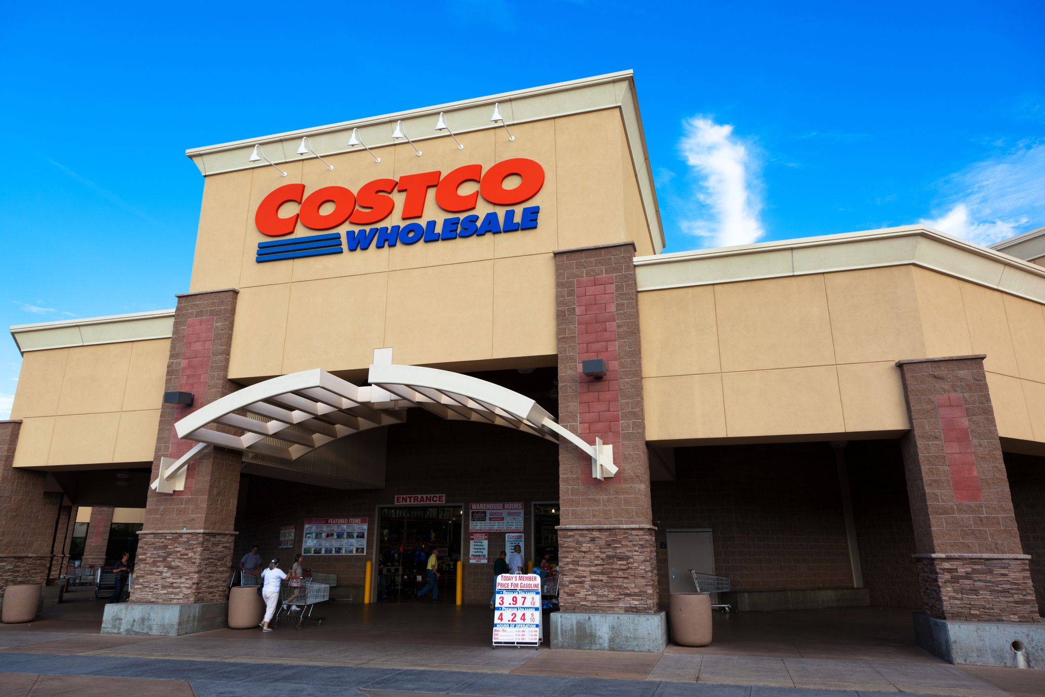 Front view of a Costco Wholesale store with a beige and red exterior, large sign above the entrance, and a few people and display signs near the entrance under a blue sky.