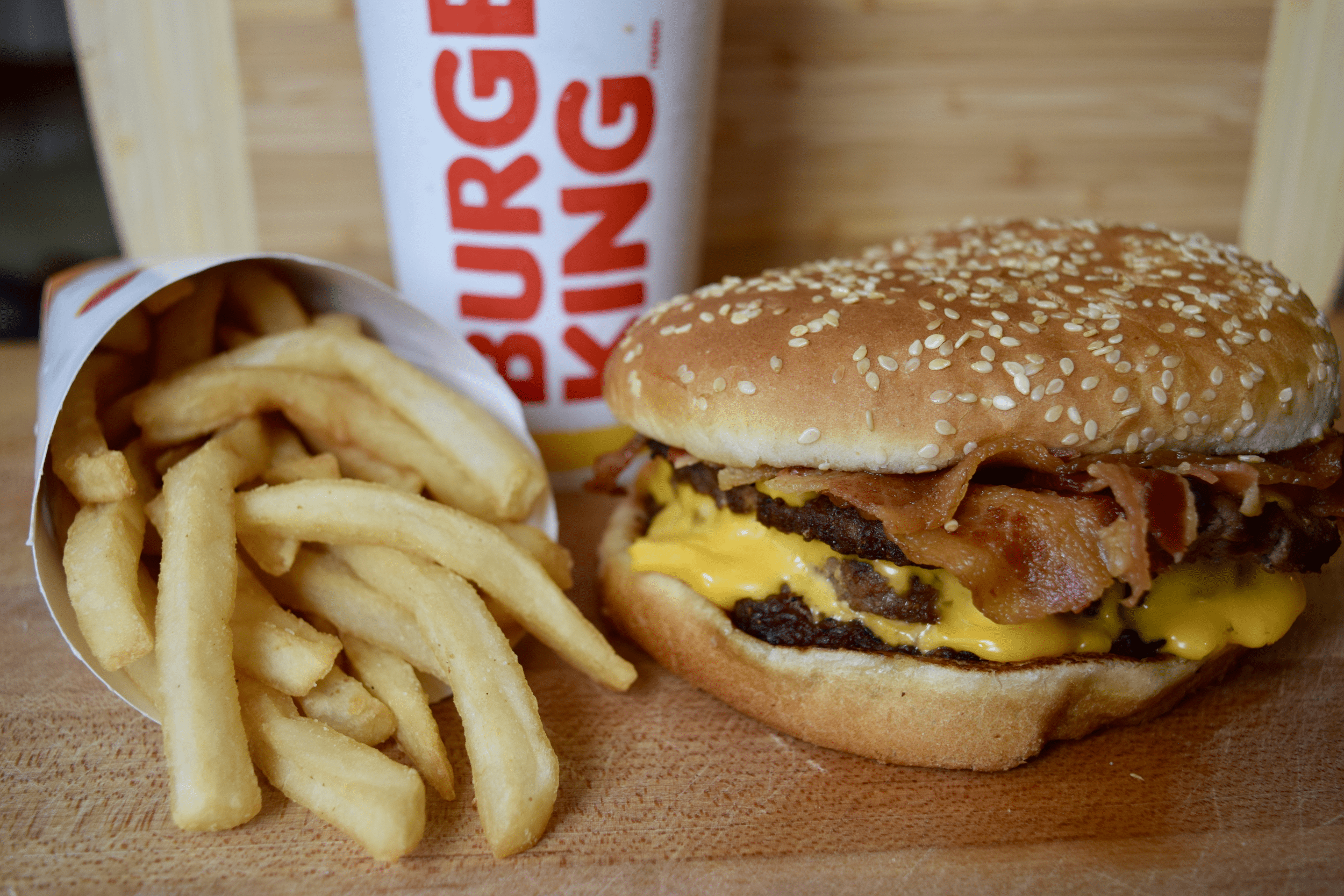 Closeup of Burger King's Triple Stacker King with fries and a drink on a wooden table with a bamboo cutting board blurred in the background