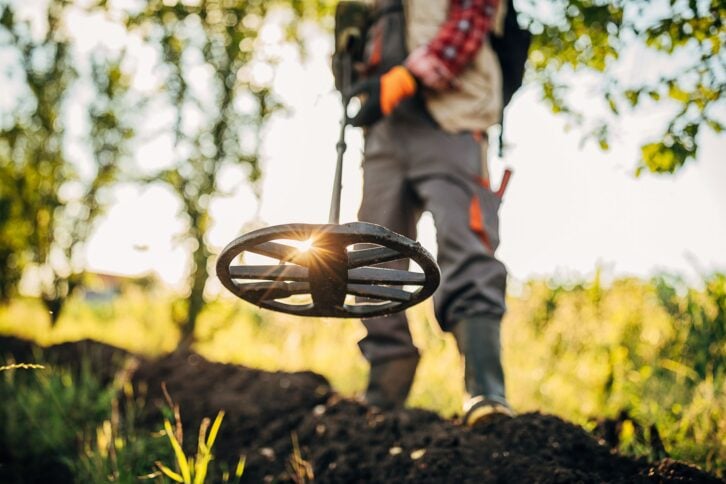 Closeup of metal detector search coil with a beam of sunlight coming through, selective focus with a man's midsection holding it, outside on a sunny day in the woods with a field in the background