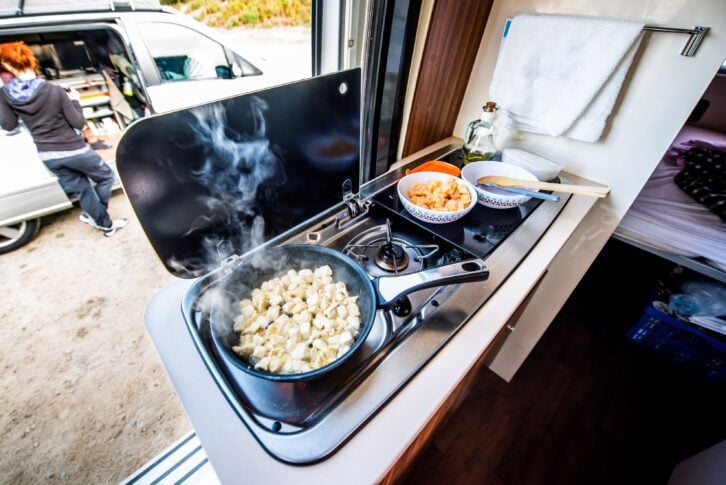 Closeup of cooking meal on stove in campervan with smoke coming from chicken in skillet, with another campervan and people in the background
