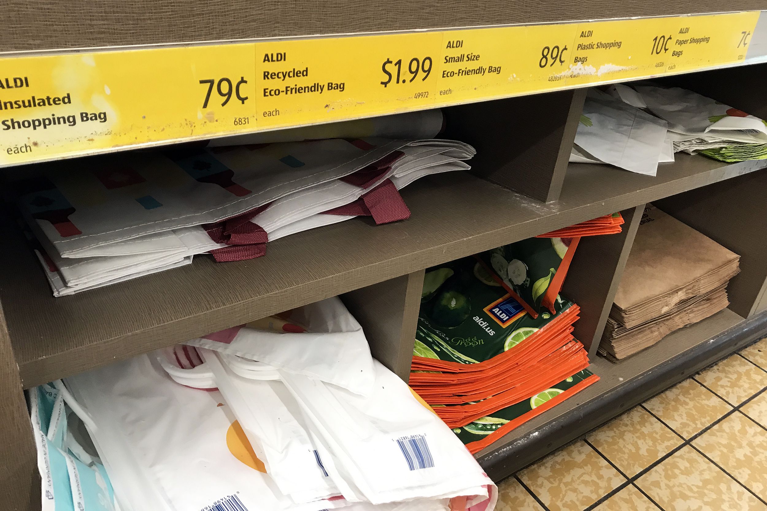 Closeup of 2 rows of cubby holes with shopping bags to purchase in an Aldi store, prices in yellow with descriptions at the top, tile floor on the right side