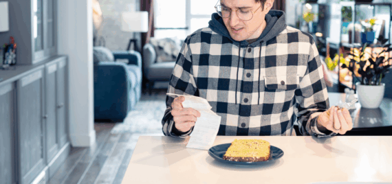 Concerned young man is looking over a receipt in one hand and holding a credit card in the other while sitting at a table in his kitchen with a pieces of bread on a blue plate, with the living room blurred in the background