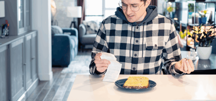 Concerned young man is looking over a receipt in one hand and holding a credit card in the other while sitting at a table in his kitchen with a pieces of bread on a blue plate, with the living room blurred in the background