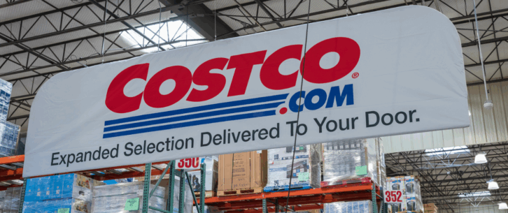 Costco.com sign hanging inside a Costco Wholesale store, Tigard, Oregon, items on the top of several aisles and ceiling with windows in the background