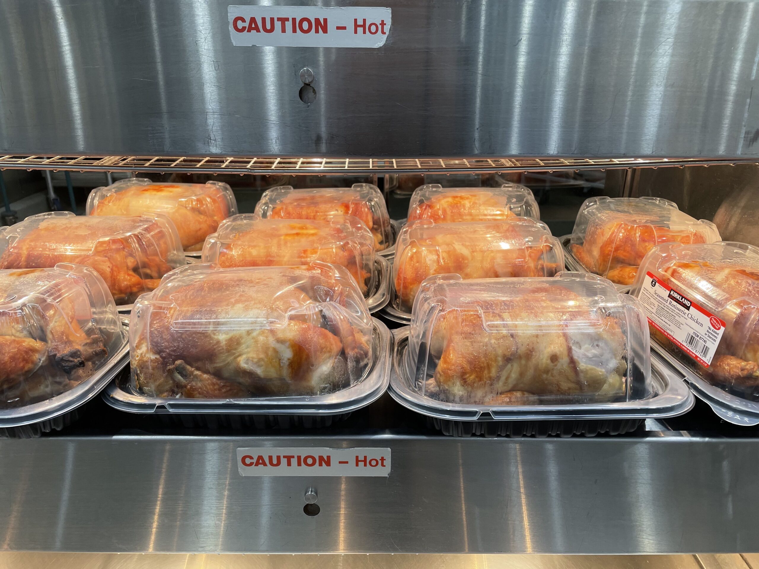Several whole rotisserie chickens in clear plastic containers are displayed on a metal warming shelf with “CAUTION – Hot” signs above and below.