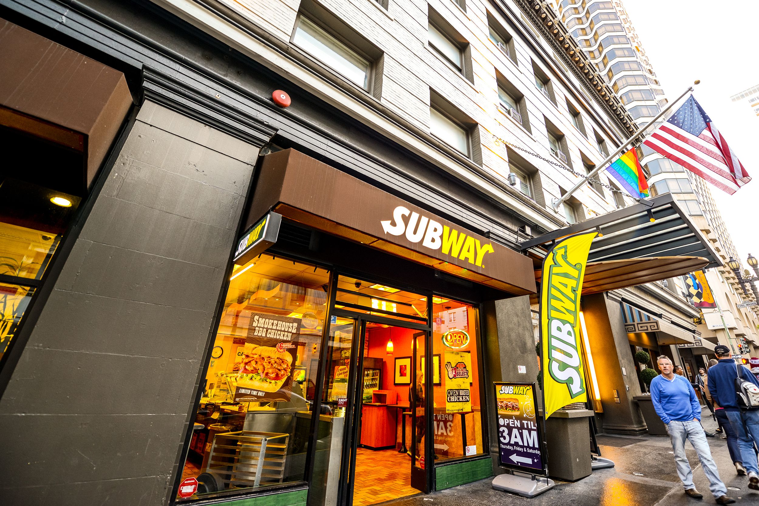 Dramatic angle view of the front exterior of a Subway restaurant, San Francisco, gay pride, Subway, and U.S. flags hanging outside, people walking along the sidewalk on a rainy day