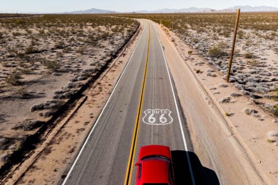 Drone view of red car driving on a lone road in the California desert on Route 66, mountains in the background