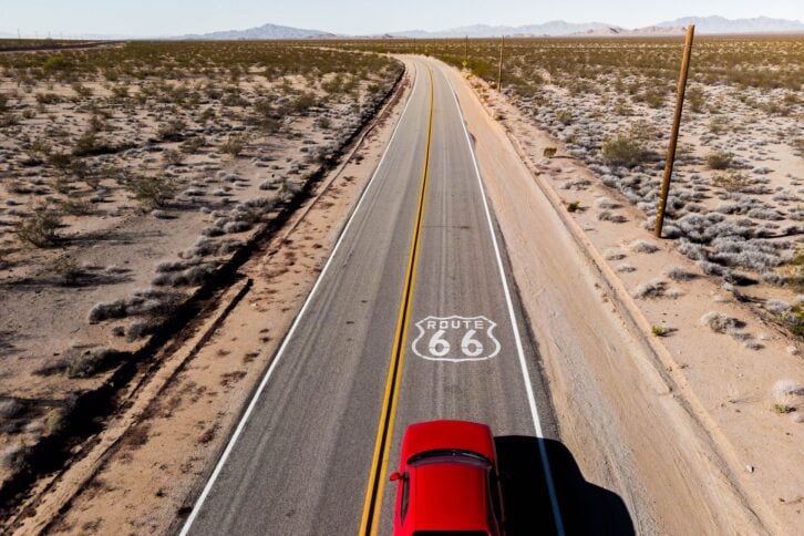 Drone view of red car driving on a lone road in the California desert on Route 66, mountains in the background