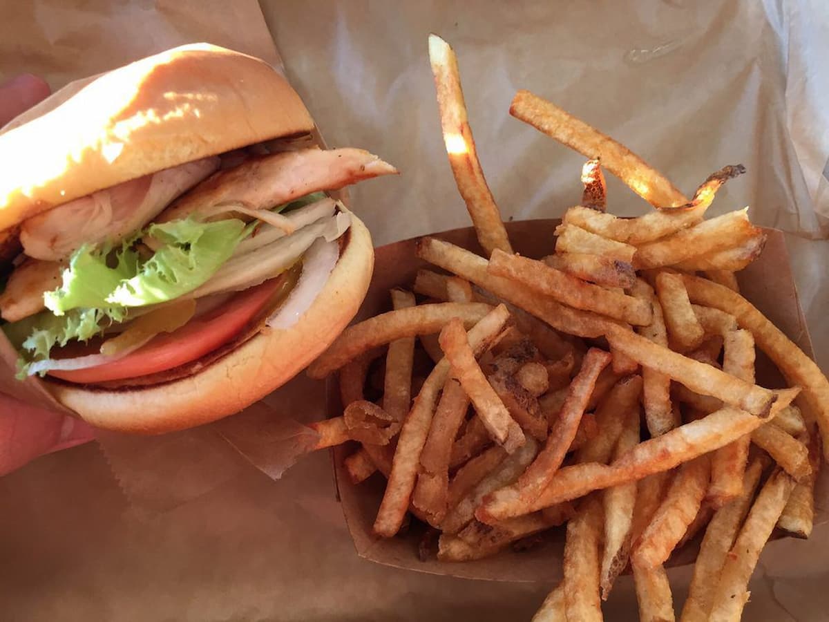 A hand holds a chicken sandwich with lettuce and tomato—like those from fast-food chains with real chicken—next to a tray of golden, crispy French fries on brown paper.