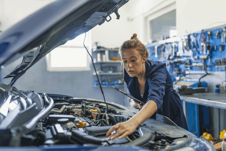 Female auto mechanic examining engine of an automobile with hood up, sideview of engine in the foreground, she is leaning over from the other side, inside a repair garage