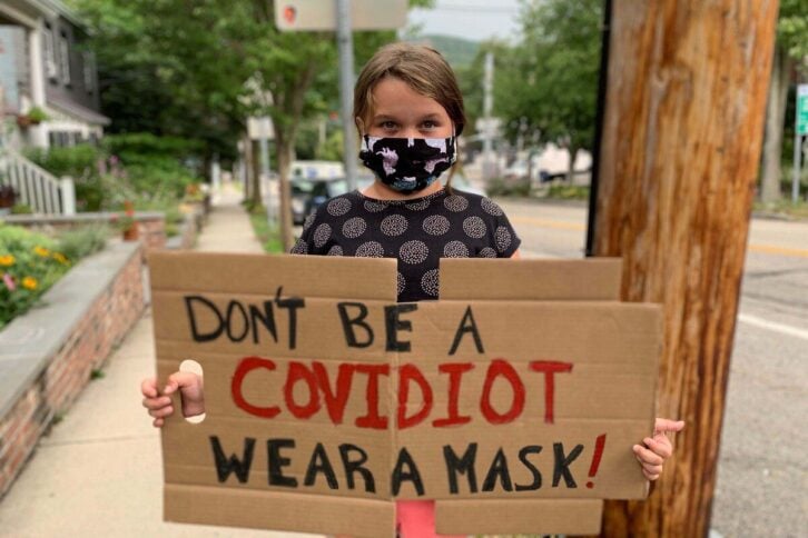 Female child holding cardboard sign that reads "Don't Be a Covidiot, Wear a Mask!", along a suberban street during a summer day