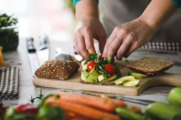 Focus on two hands putting avocado, tomatoes, and herbs on a slice of bread for a sandwich, on a wooden cutting with more slices of bread, on a wooden table, ingredients and vegetables blurred in the foreground and on sides, window in background blurred