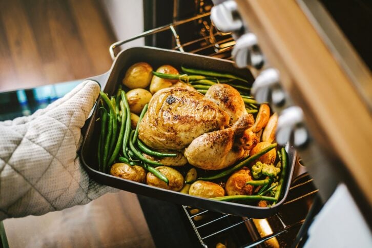 Focus on a whole chicken with potatoes and green beans in a pan out of the oven, one hand wearing an oven mitt holding it, with a blurred background of the range and wooden floor