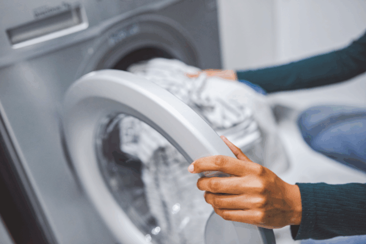 Focus on woman putting clothes into a front load washing machine, her hand on the door, selective focus, washing machine blurred in the background