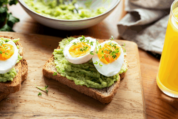 Focus on a slice of avocado toast with soft boiled eggs, next to a pile of same, on a wooden cutting board next to a glass of orange juice, with a blurred background of ingredients and napkins