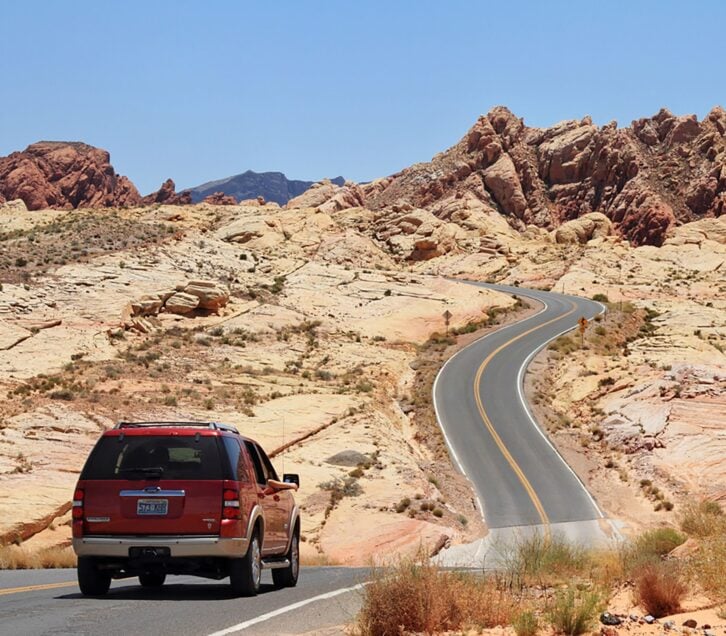 A Ford Explorer SUV travels on a winding desert road toward distant red rock formations in the Valley of Fire state park in Nevada.
