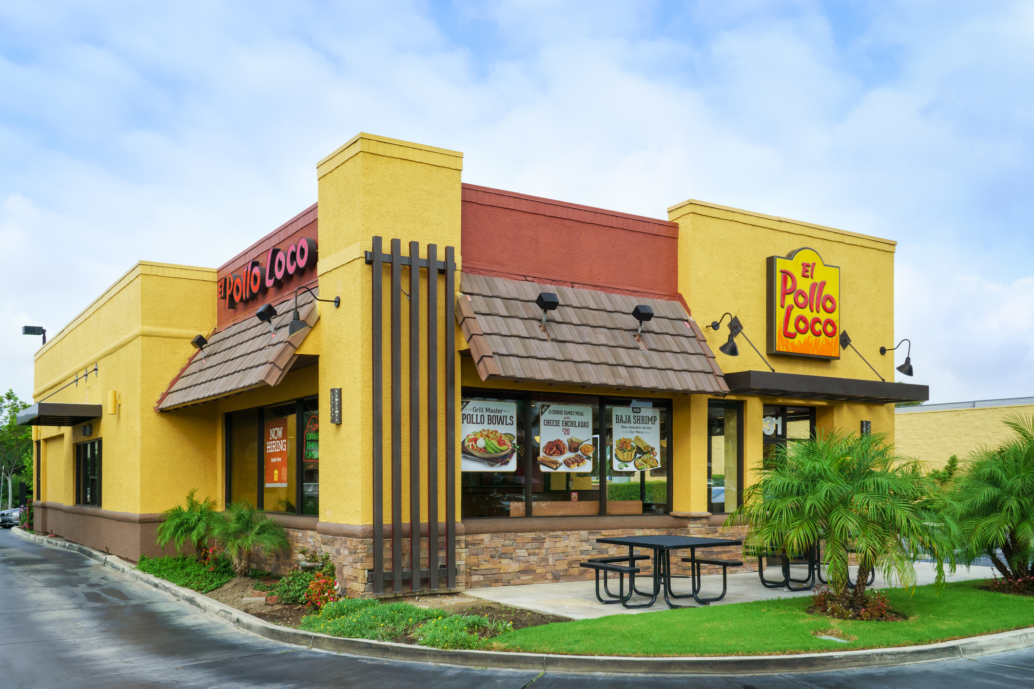 Front exterior of El Pollo Loco restaurant, Costa Mesa, California, greenery with seating area and drive-thru to the left, against a blue sky with white clouds