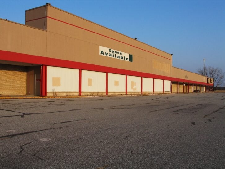 Front exterior of an empty, abandoned store in a strip mall with windows boarded up, 'space available' sign on top, and street in the foreground, against a blue sky with bare trees to the right